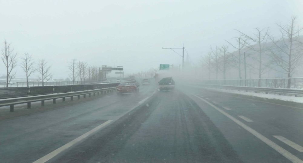 Snow blowing off a truck traveling ahead · Snow is falling, so the road is slippery