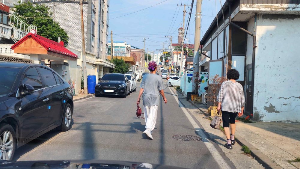 Piyodalar yo'lagi va qatnov qismi ajratilmagan, markaziy chizig'i yo'q yo'l · 2 nafar piyoda ketmoqda · Yo'lning chap tomonida avtomobillar turibdi
