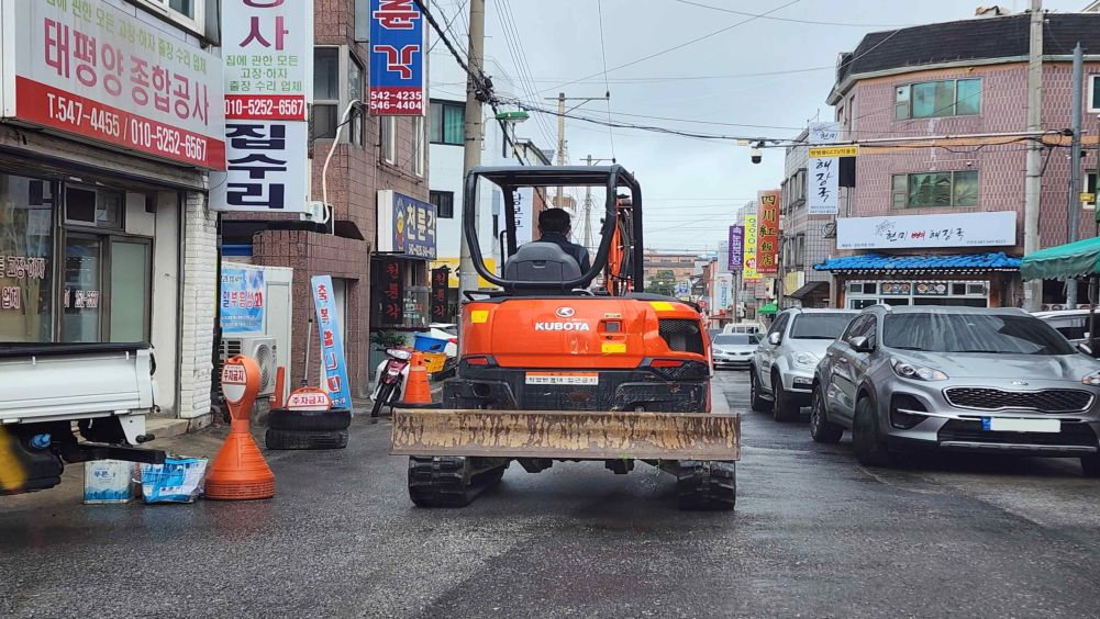 Đường hẻm khu dân cư không phân chia vỉa hè và phần đường xe chạy · Mặt đường ướt do mưa · Máy xây dựng phía trước đang chạy chậm · Bên phải đường có các xe đang đỗ