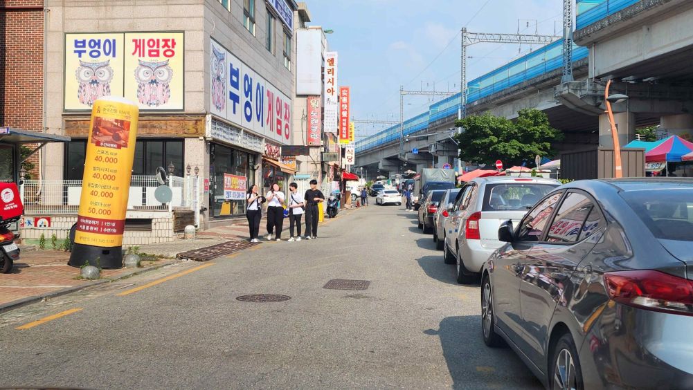 Back street around shops with no separation between sidewalk and roadway · Cars are parked in a row on the right side of the road · Pedestrians are walking toward you on the left side of the road · There are steel manhole covers here and there on the road