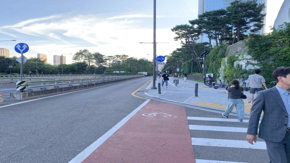ทางแยกรูปตัวที (รูปตัว ㅏ) · มีทางม้าลายและทางข้ามจักรยาน · คนเดินเท้ากำลังข้ามทางม้าลาย · มีป้ายทางร่วมจักรยานและคนเดินเท้าอยู่ข้างหน้า · ผู้ขี่จักรยานกำลังเข้ามาบนทางร่วมจักรยานและคนเดินเท้า